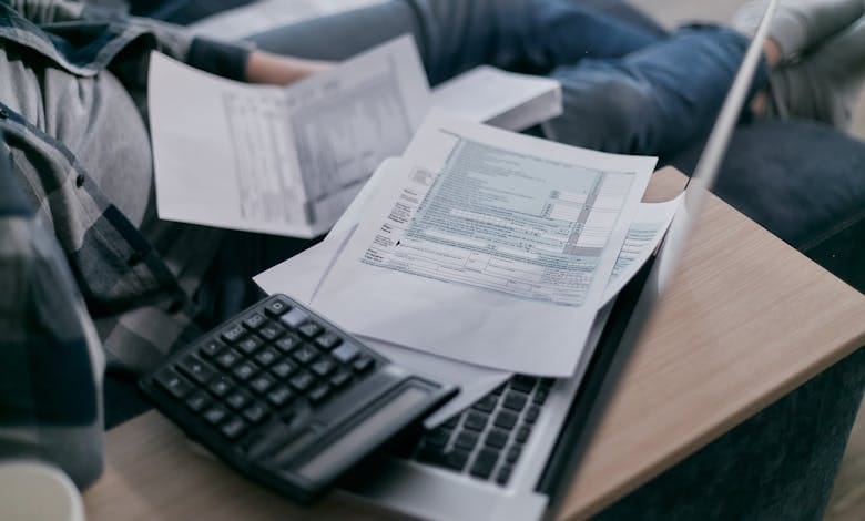 A young adult using a calculator and laptop to manage finances at home, surrounded by papers.